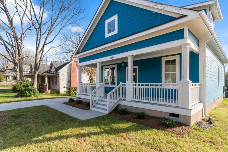 Cozy blue suburban house with a white front porch surrounded by lush greenery on a clear day.