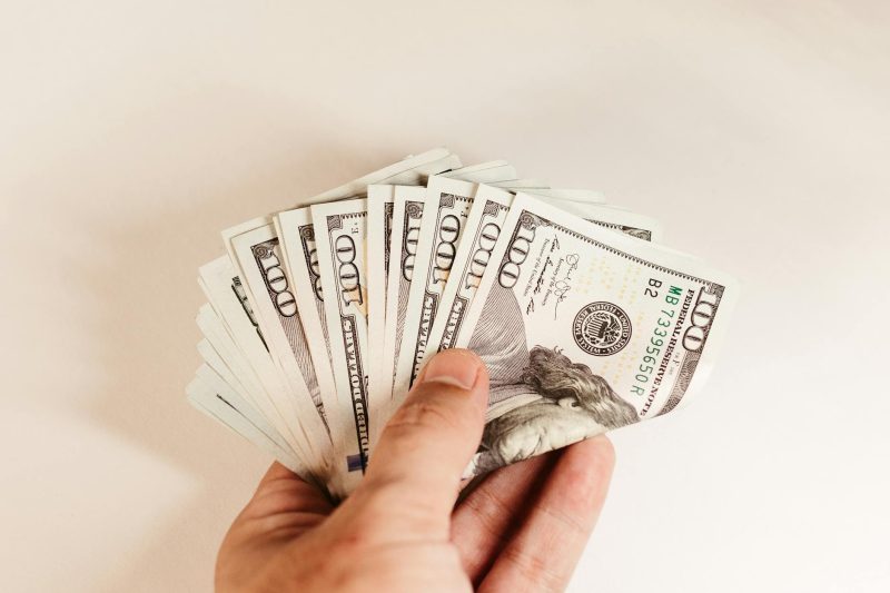 Close-up of a hand holding folded US hundred-dollar bills against a white background.