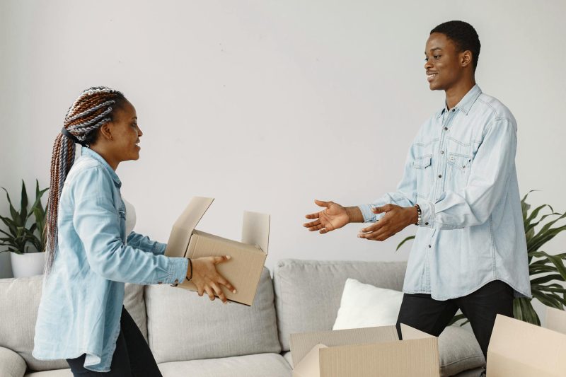 A young couple joyfully carrying cardboard boxes into a bright, newly furnished living room.