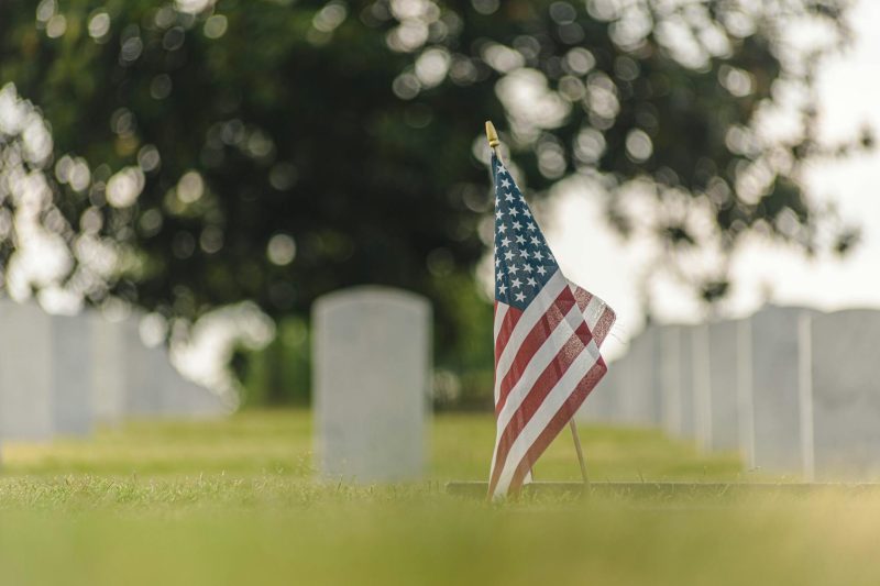American flag standing tall at a cemetery with blurred headstones in the background.
