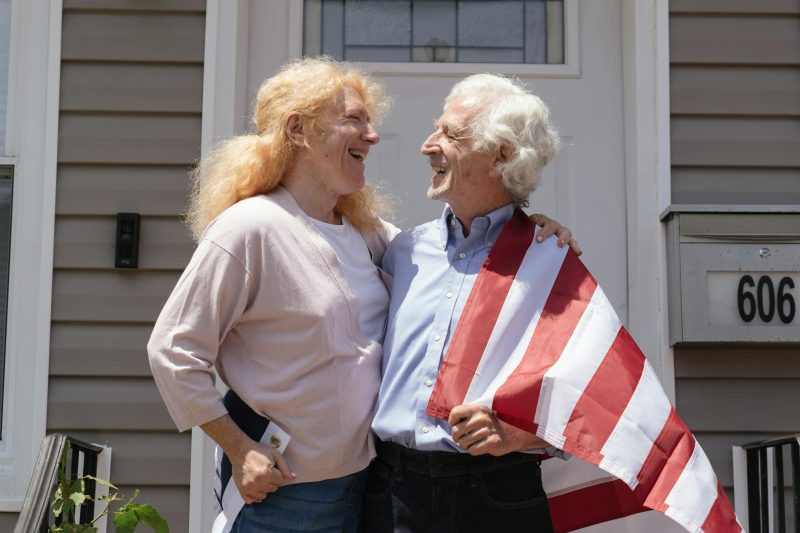 Happy senior couple stands outdoors, embracing with an American flag in the background.